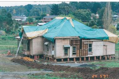 Weatherboard house sitting on exposed stumps, roof covered with tarpaulins, parts of walls missing.