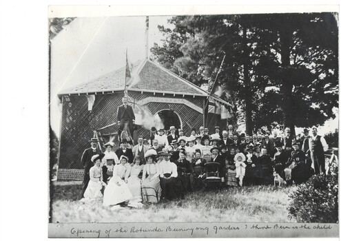 Crowd of people in Victorian dress, infront of wooden lattice rotunda, large cedars in background.