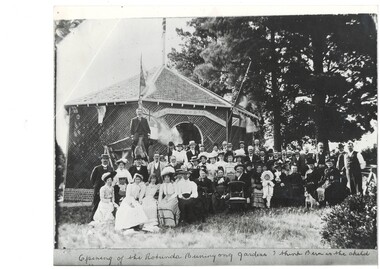 Crowd of people in Victorian dress, infront of wooden lattice rotunda, large cedars in background.