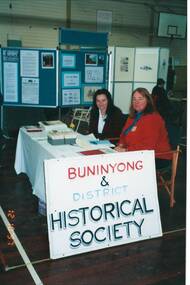 Display table covered by sheet, display board , two ladies sitting.