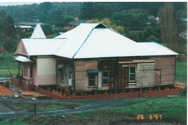 View of weatherboard house being relocated, on raised stumps in cleared area of block,  some walls removed.