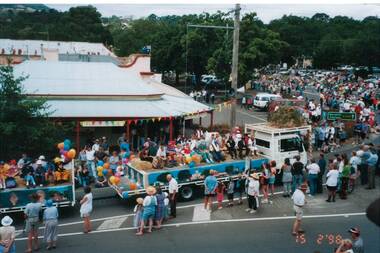 Looking down on procession past shop, double tray float with schoolchildren passing, crowds lining streets.