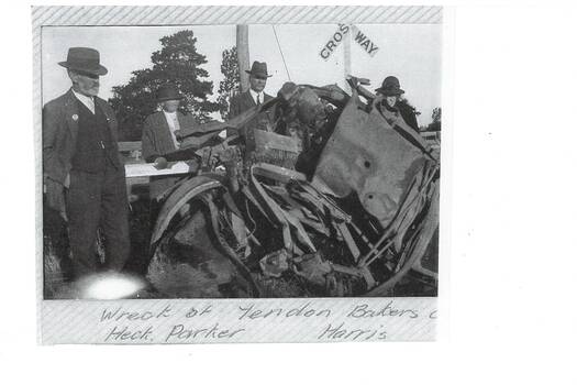 Four men in pre-WWII style suits and hats, standing around mangled wreck of a vehicle, railway crossing sign behind them.