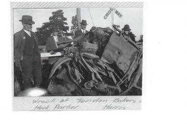 Four men in pre-WWII style suits and hats, standing around mangled wreck of a vehicle, railway crossing sign behind them.