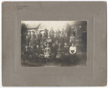 1920s primary school grade photo, three rows mixed boys and girls, winter day-clothes, in front of school building.