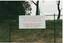 White notice board, red lettering, hung on chain link fence, grass and clay bank and trees behind.