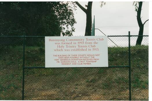 White notice board, red lettering, hung on chain link fence, grass and clay bank and trees behind.