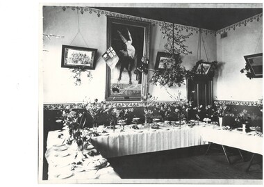 Interior of supper room showing cloth covered tables set with vases of flowers and cakes 