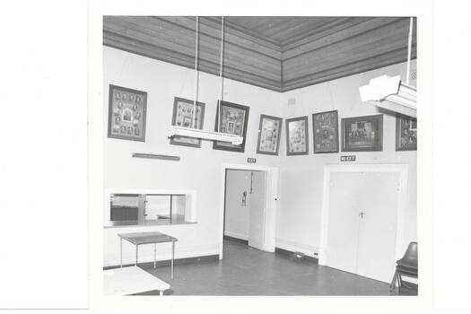 Corner of large, white painted room, high wooden ceiling, kitchen servery on left, single door centre, double doors on tight, large framed photographs around walls above door height./