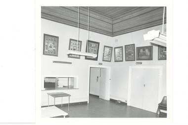 Corner of large, white painted room, high wooden ceiling, kitchen servery on left, single door centre, double doors on tight, large framed photographs around walls above door height./
