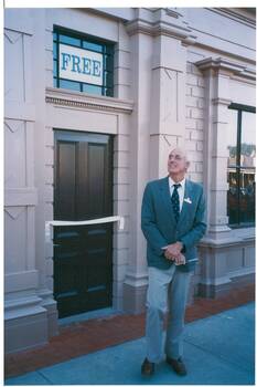 Gentleman in blazer and tie, holding a pair of scissors, outside decorated wooden façade of old building, ribbon across the doorway.