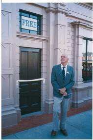 Gentleman in blazer and tie, holding a pair of scissors, outside decorated wooden façade of old building, ribbon across the doorway.