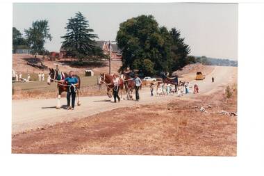 Procession of horse-drawn vehicles along dirt road, school in the distance.