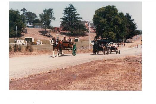 Two horse-drawn carts on dirt road, school in distance.