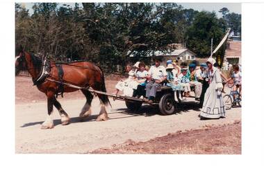 Horse-drawn dray, packed with children, lady in Victorian costume, on a dirt road.