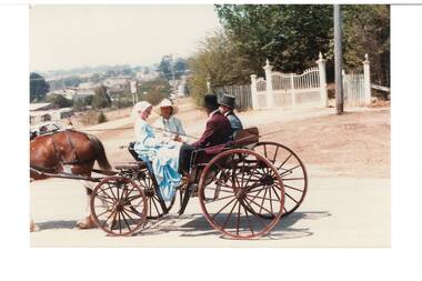 Two ladies and two gentlemen in Victorian dress, seated in horse-drawn buggy, at intersection of two dirt roads, ornate white iron gates behind them.