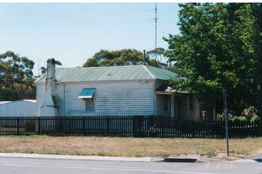 Side view of C19th weatherboard cottage, white, chimney at rear, veranda along front, low black picket fence,
