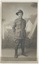 An Unknown World War 1 Sergeant stands in uniform in front of a photographic studio backdrop featuring military tents