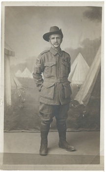An Unknown World War 1 Sergeant stands in uniform in front of a photographic studio backdrop featuring military tents