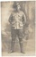 A black and white image of a World War 1 Australian Soldier in uniform standing at ease in front of a photographic studio backdrop showing a stylized entryway featuring plants and stairs 