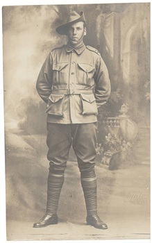 A black and white image of a World War 1 Australian Soldier in uniform standing at ease in front of a photographic studio backdrop showing a stylized entryway featuring plants and stairs 