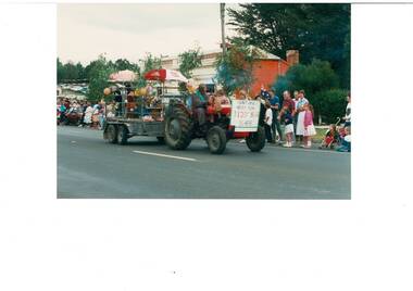 Tractor pulling a trailer, driver in air-man's gear, sign on front, people on the side of the road, building in the  background.