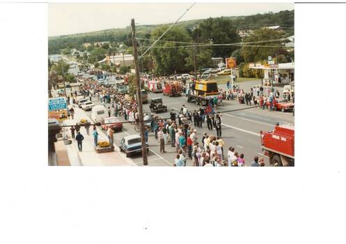 Looking down on procession of trucks and floats passing a service station, people lined up either side of the road.
