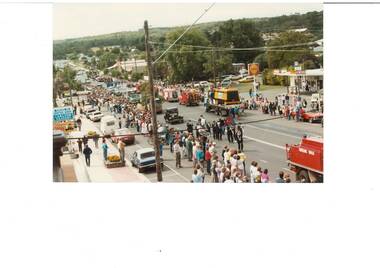 Looking down on procession of trucks and floats passing a service station, people lined up either side of the road.
