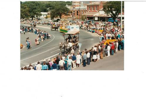 Looking down at procession, horse-drawn carts, crowds lining sides of the road, old brick buildings in thje background.