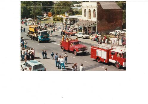 Looking down on procession passing old two-story brick shop and service station, led by two fire trucks./