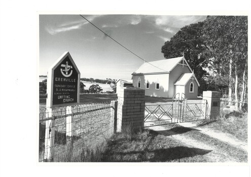 White wooden church, gothic shaped windows, trees to the right, geometric iron gate in brick pillars and wire fence in foreground.