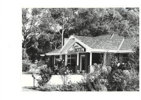 Monochrome photo, hotel, low building, tiled roof, tall trees behind, driveway and rose gardens in foreground.