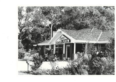 Monochrome photo, hotel, low building, tiled roof, tall trees behind, driveway and rose gardens in foreground.
