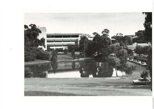 B&W photograph, looking across ornamental lake at 4-story modern building, verandas right across front, all levels, trees and lawn surrounding lake.