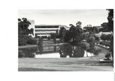 B&W photograph, looking across ornamental lake at 4-story modern building, verandas right across front, all levels, trees and lawn surrounding lake.