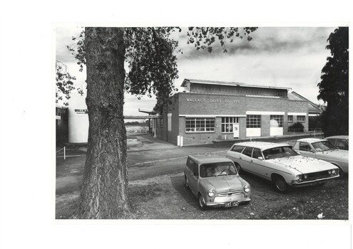 Brick factory, metal framed windows, cars parked under trees in foreground, to the left big milk vat.