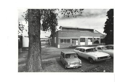 Brick factory, metal framed windows, cars parked under trees in foreground, to the left big milk vat.