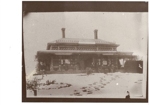 Elevated view of impressive, single story brick victorian house, deep veranda on all sides, bay windows, tall, ornate brick chimneys.  The ground is covered in snow, with footprints going towards the front door.