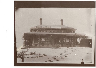 Elevated view of impressive, single story brick victorian house, deep veranda on all sides, bay windows, tall, ornate brick chimneys.  The ground is covered in snow, with footprints going towards the front door.
