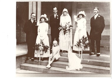 Wedding party , 1930's clothing, standing on church steps. Three couples, bride and groom centre, groomsman and bridesmaid either side, flower girl in front. Men in morning suits, women in white gowns, flower girl in white frock sitting on steps.