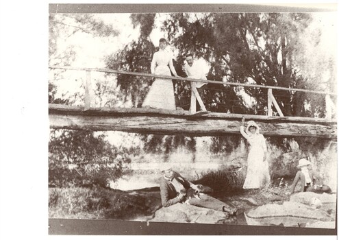 Wooden bridge near small lake, lady and man on bridge, man and woman reclining on rocks below the bridge with another lady in between.  All are in Edwardian summer clothes.