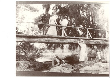 Wooden bridge near small lake, lady and man on bridge, man and woman reclining on rocks below the bridge with another lady in between.  All are in Edwardian summer clothes.