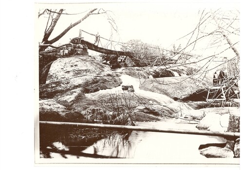 A black and white image of a creek featuring a small water wheel in the foreground and Arthur Fisken standing on a wooden bridge to the rear.