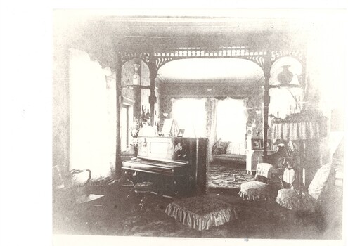 House interior, showing period furnishings, piano with stool, chairs, screens, and an ornate lampshade.  