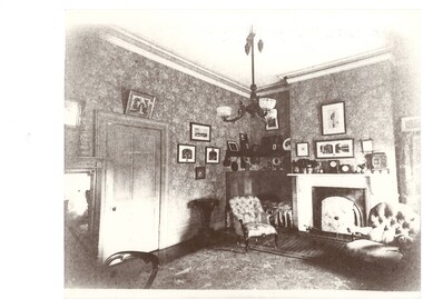 Black and white interior view of the drawing room Of Lal Lal Park featuring furniture,  decorative pictures and ornaments around the fireplace, an elaborate ceiling light, cornice work and wallpaper.