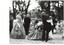 Circle of dancers in period dress, C19th, holding hands, outdoors, trees in background. 