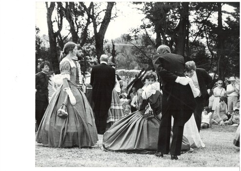 Circle of dancers in period dress, C19th, holding hands, outdoors, trees in background. 