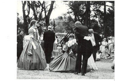 Circle of dancers in period dress, C19th, holding hands, outdoors, trees in background. 