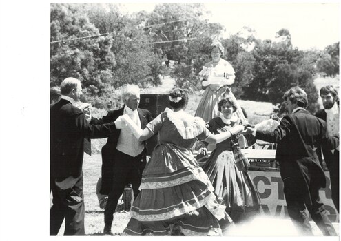 Circle of dancers in period dress, C19th, holding hands, outdoors, trees in background. Another lady in similar costume stands on a platform behind them.