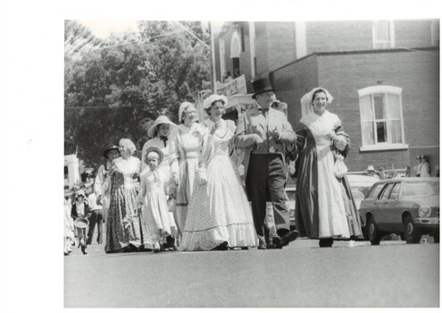 Group of woman and girls in C19th period dress, led by two women arm in arm with a gentleman in C19th frock coat and top hat, walking past a brick building.